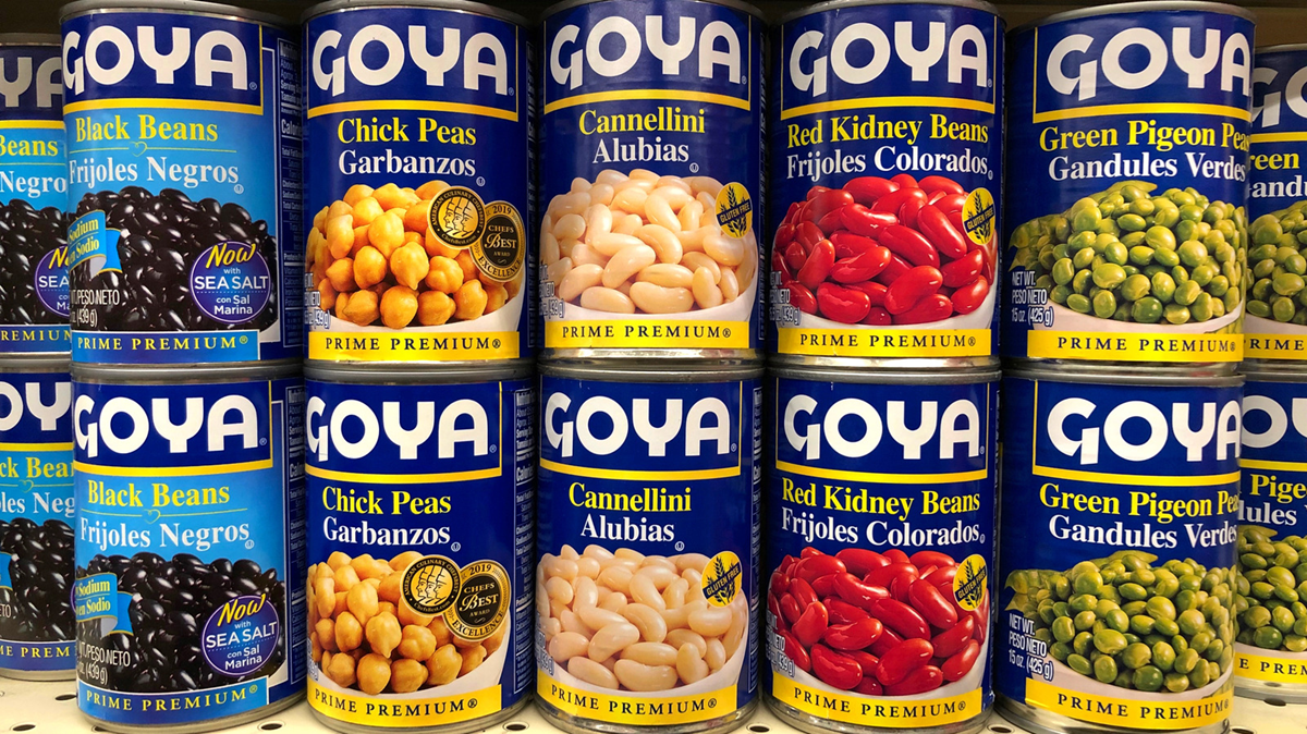 Cans of Goya beans, including black beans, chickpeas, cannellini, red kidney beans, and green pigeon peas, are stacked side by side on a grocery store shelf.