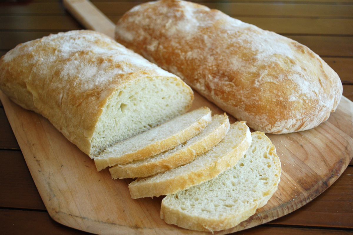 Two loaves of rustic bread on a wooden cutting board; one loaf is whole and the other is partially sliced into several pieces. The bread has a golden, flour-dusted crust and a soft, airy interior.