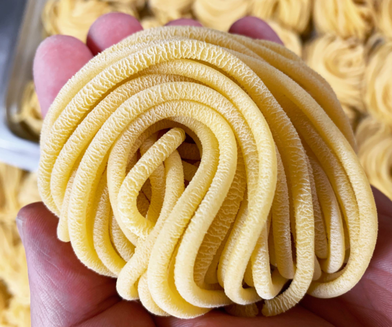 A close-up of a hand holding a nest of uncooked yellow pasta noodles, showing their soft texture and tightly coiled, ribbon-like shape. Other pasta nests are visible in the blurred background.