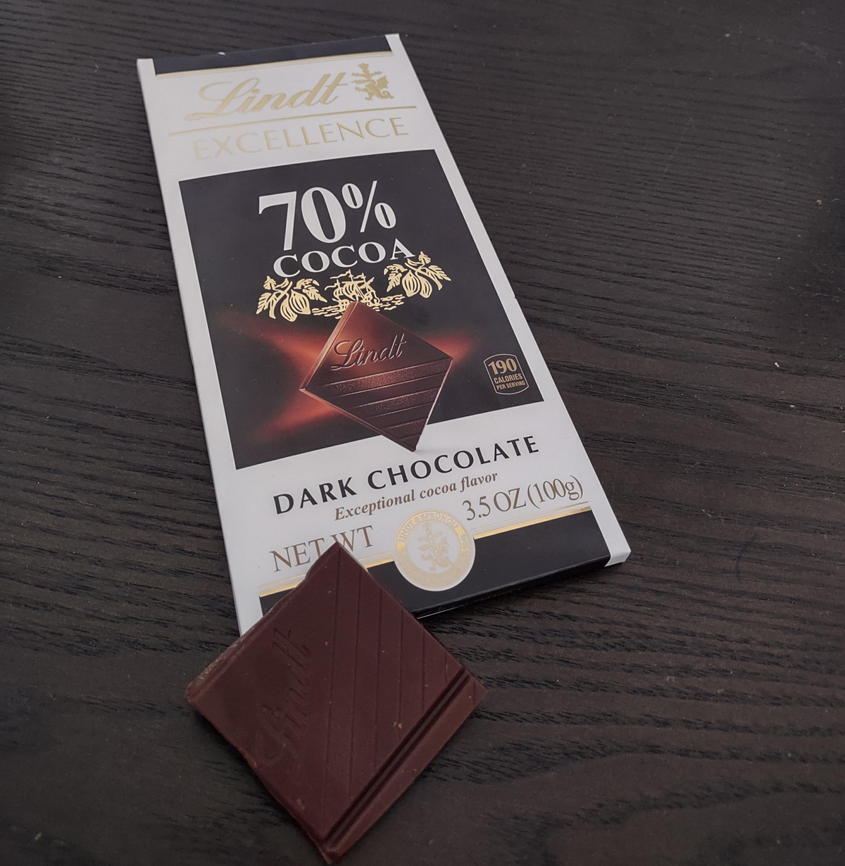 A Lindt Excellence 70% Cocoa Dark Chocolate bar, partially unwrapped on a dark wooden surface, with two squares of chocolate placed in front of the packaging.