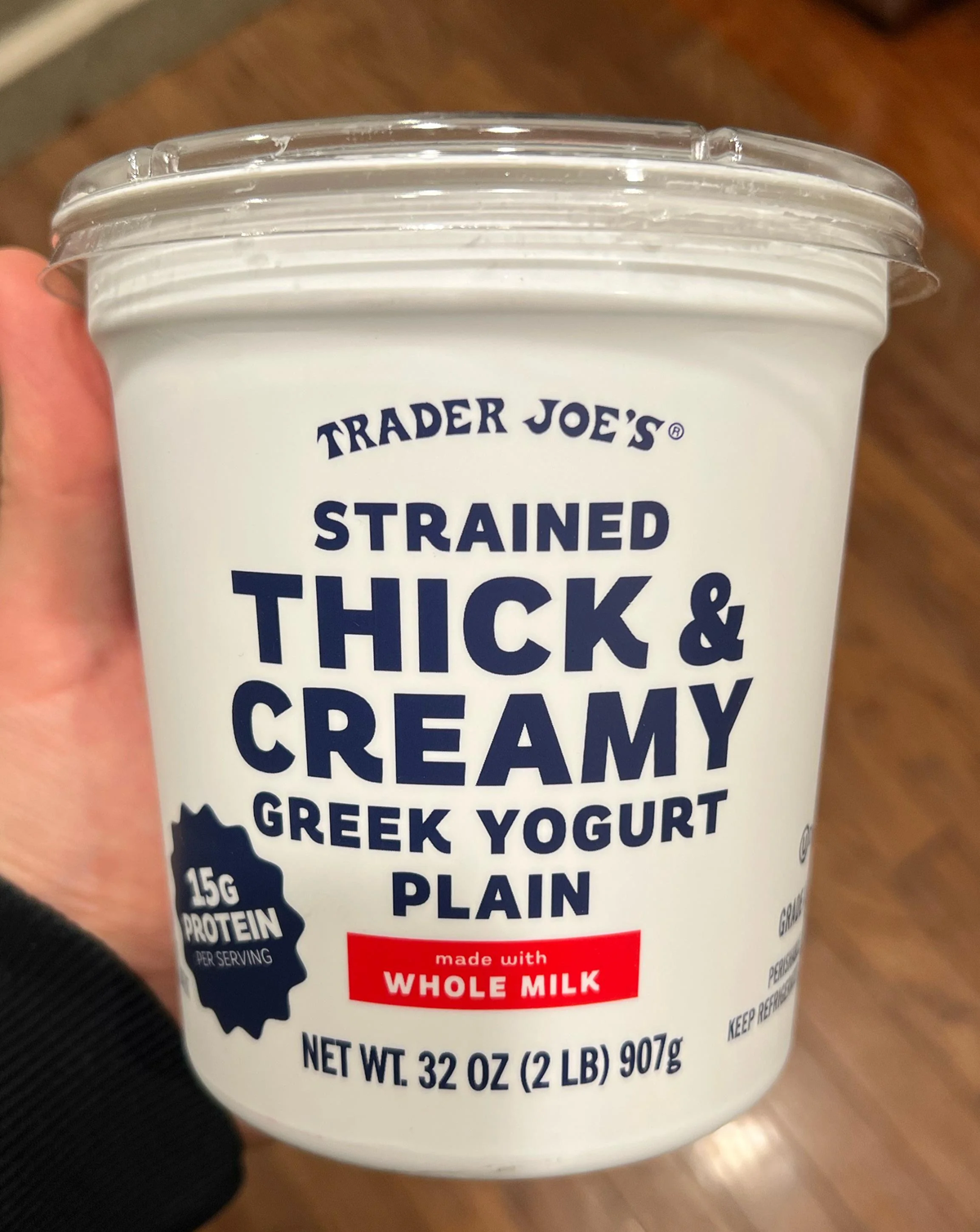 A hand holds a 32-ounce container of Trader Joe's Strained Thick & Creamy Plain Greek Yogurt, made with whole milk. The packaging highlights 15g protein per serving. The background is a wooden floor.