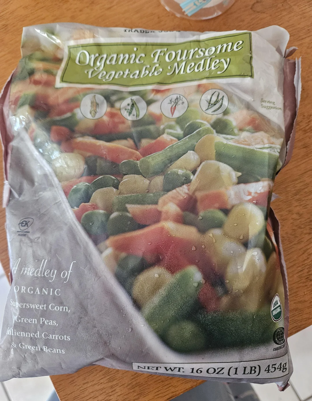 A bag of Organic Foursome Vegetable Medley featuring a mix of sweet corn, green peas, carrots, and green beans. The bag weighs 16 oz (1 lb) and is placed on a wooden surface.