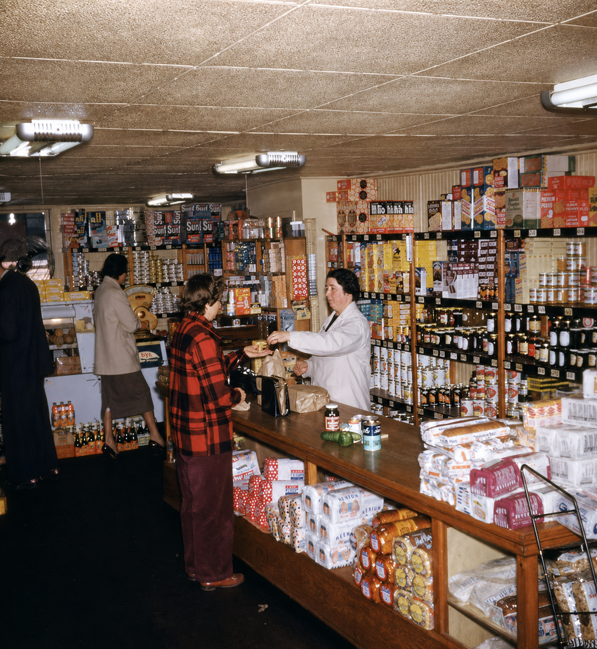 A grocery store scene with shelves full of canned goods, jars, and packaged foods. A woman behind the counter serves a customer in a red plaid coat while other shoppers browse. The store has a cozy, vintage feel. Vintage Photos Grocery Store 1950s