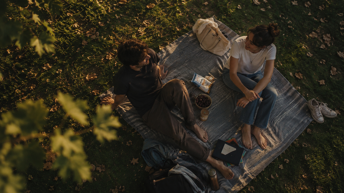 A man and woman sit barefoot on a blanket having a picnic in a grassy park, with snacks, drinks, a book, and bags around them. They are talking and surrounded by dappled sunlight and fallen leaves.