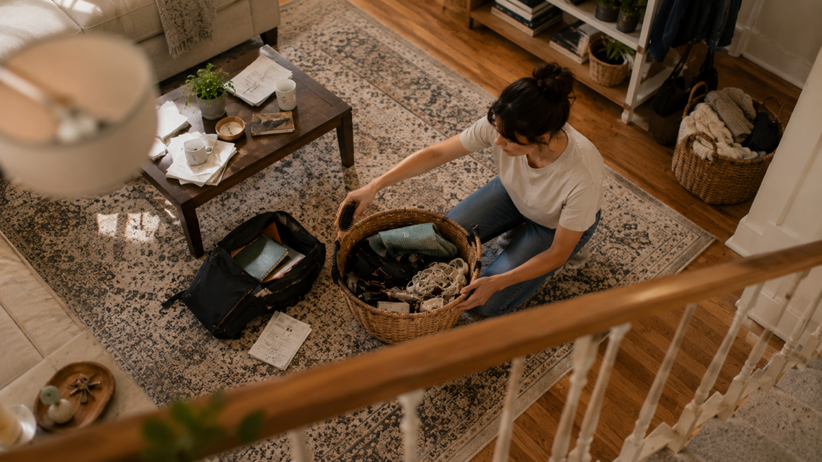 A woman sits on a patterned rug in a living room, organizing items into a wicker basket. An open bag, notebooks, and papers are nearby. A coffee table with various objects is in front of her.