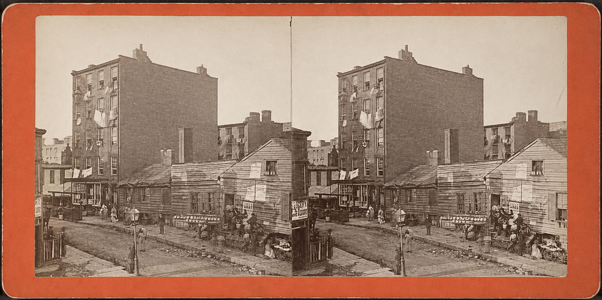 A sepia stereograph shows a city street with wooden houses in front of taller brick apartment buildings; people stand and gather near the houses, with laundry hanging from the windows above.
