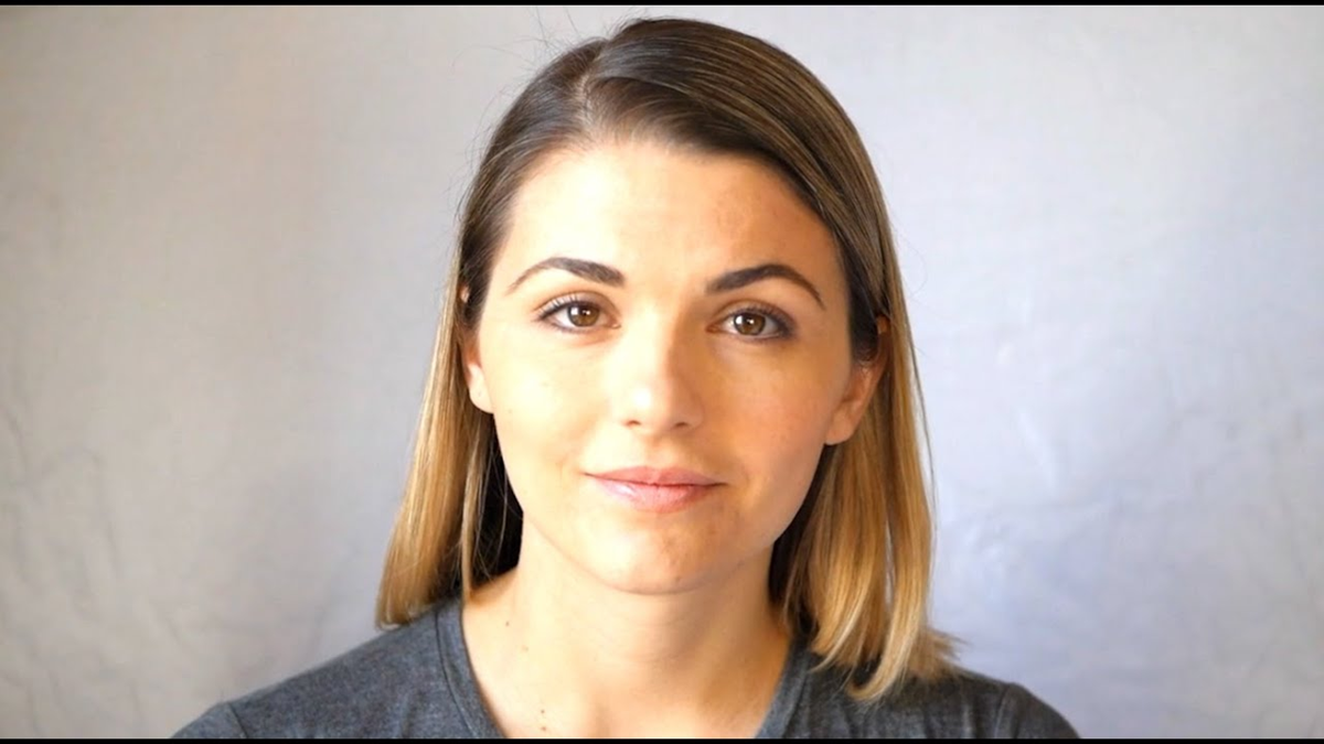 A woman with straight, shoulder-length blonde hair and natural makeup is facing the camera and smiling softly. She is wearing a grey shirt and is posed against a plain, light background.