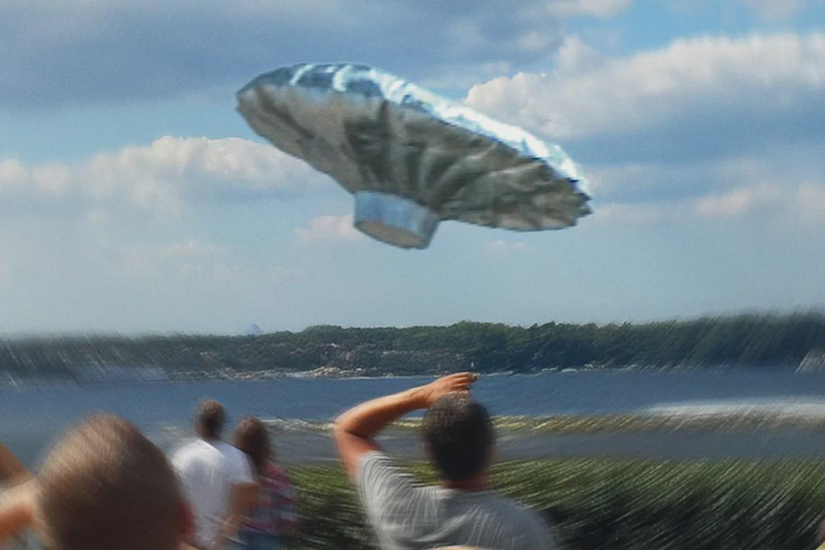 A group of people outdoors look up at a shiny, balloon-like object shaped like a flying saucer floating in the sky near a body of water, with trees and clouds in the background.