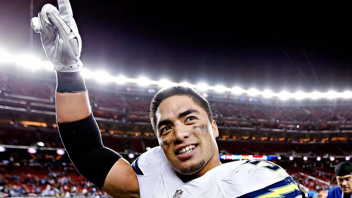 A football player in uniform smiles and points upward with his right hand on a brightly lit stadium field at night, with cheering fans visible in the background.