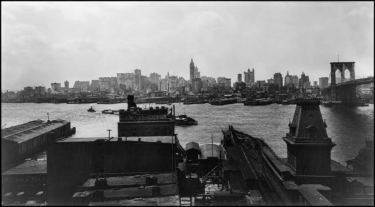 Black and white photo of the New York City skyline in the early 1900s, showing the East River, boats, industrial rooftops, and the Brooklyn Bridge on the right side of the image.
