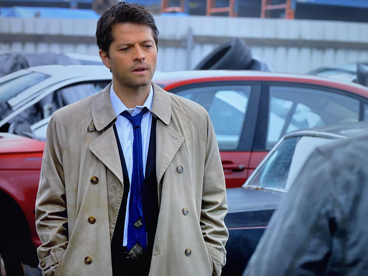 A man wearing a beige trench coat, blue tie, and dress shirt stands outdoors in a parking lot with cars and tires in the background, looking slightly to the side with his hands in his pockets.