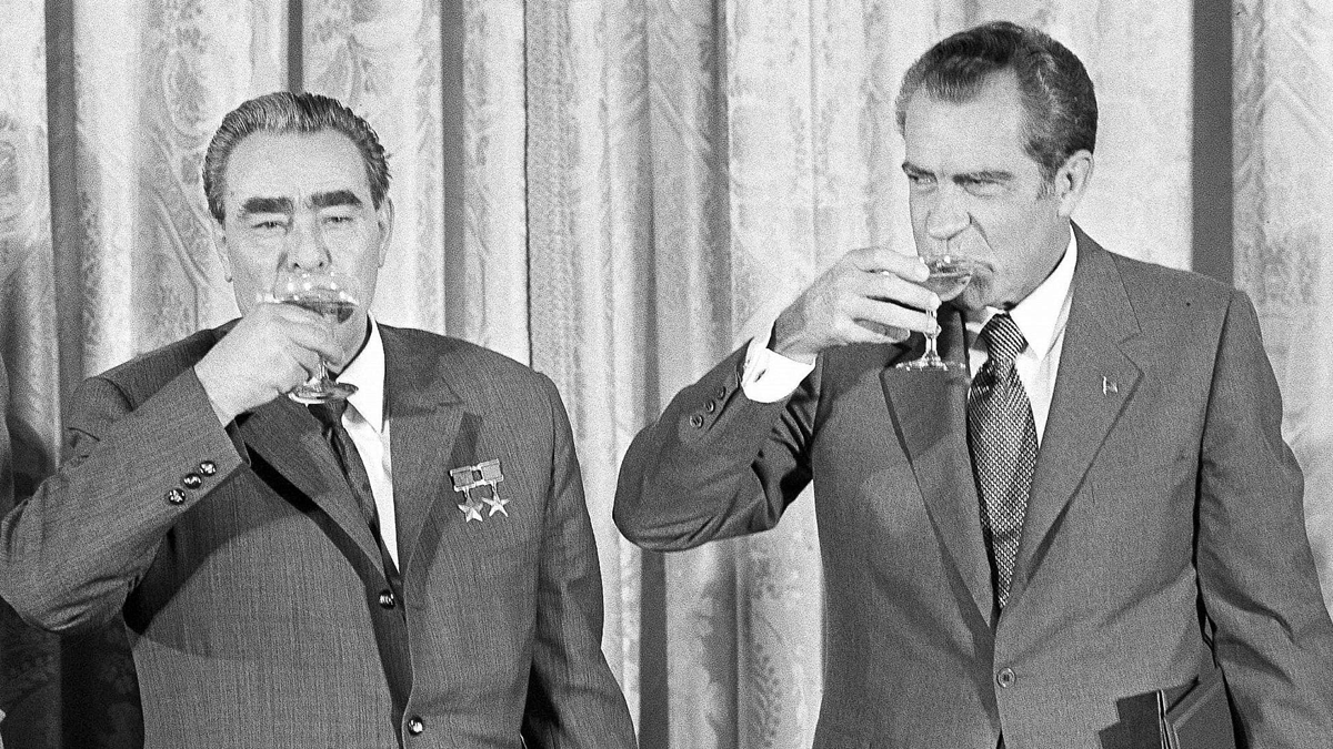 Two men in suits stand side by side, each holding a glass and sipping a drink. One man wears medals on his jacket. A patterned curtain hangs in the background. The image is black and white.