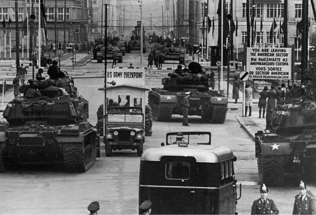 Black and white photo of US tanks and military vehicles at Checkpoint Charlie in Berlin, with soldiers and signs indicating the border between American and Soviet sectors during the Cold War.