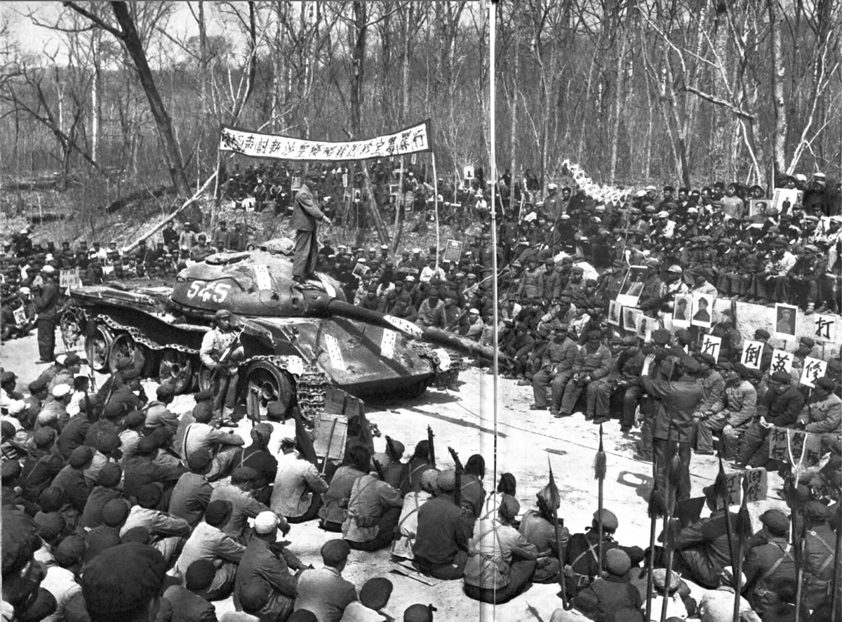 A large crowd of people, many in uniform, gather outdoors around a tank with the number 545. Banners with Chinese characters hang above as leafless trees line the background, suggesting a public assembly or event.