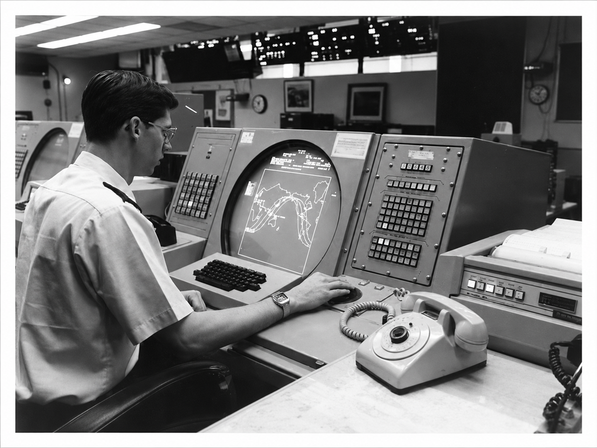 A man sits at a vintage control center console, looking at a radar screen displaying maps and data, surrounded by buttons and an old-fashioned rotary telephone.