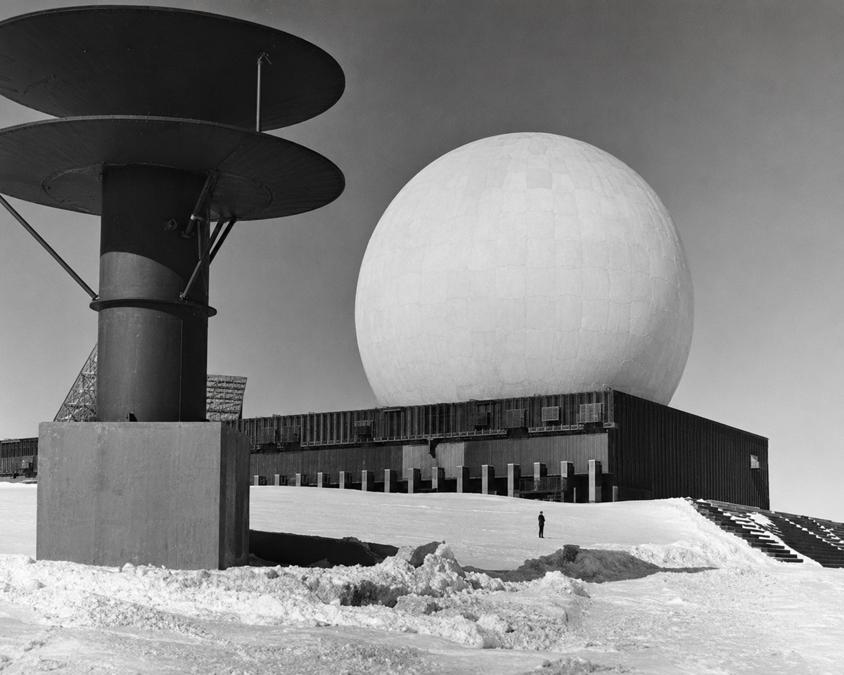 A person stands in snow near a large spherical radar dome atop a rectangular building, with a tall antenna structure in the foreground, under a clear sky.