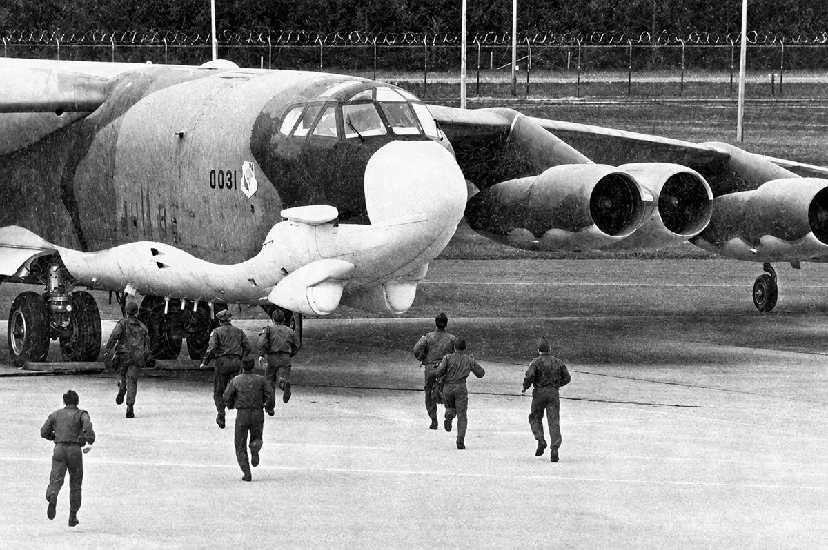 A group of military personnel in uniform runs toward a large bomber aircraft parked on a runway, with trees and a fence visible in the background.