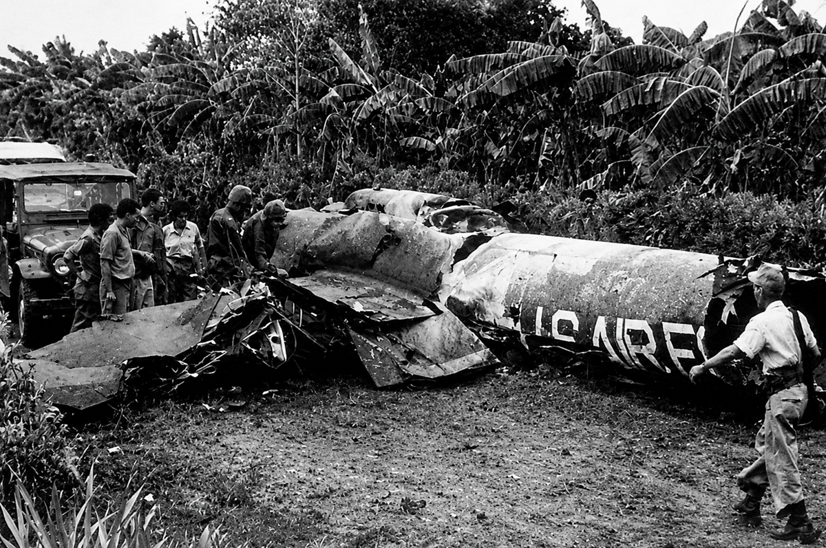 A group of people inspects the wreckage of a U.S. Air Force aircraft in a grassy, forested area, with dense foliage and a military vehicle visible in the background.