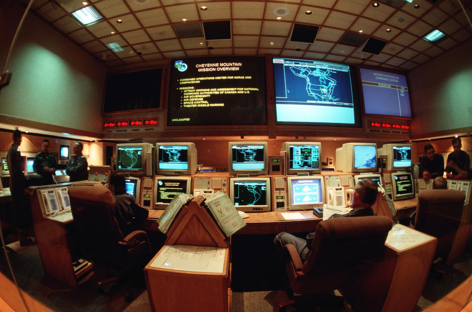 A control room with people seated at consoles, multiple computer monitors displaying maps and data, and large screens on the wall showing mission overviews and information.