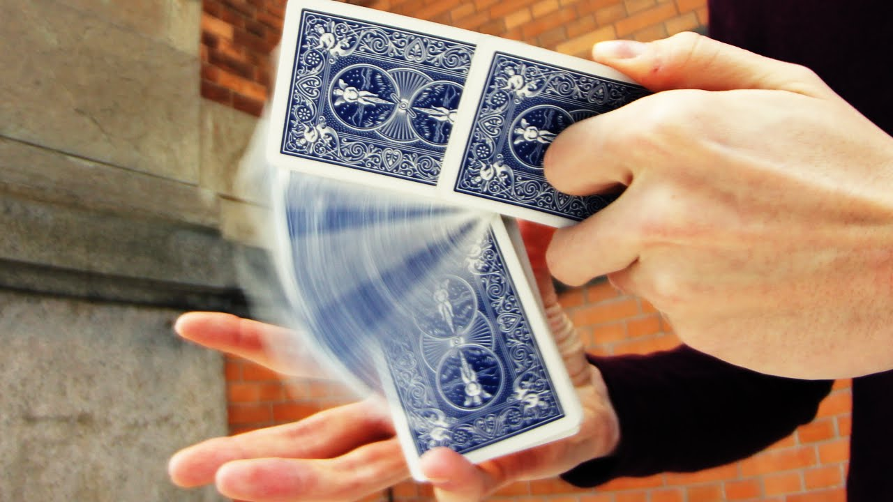 A person skillfully shuffles playing cards in midair, performing a cardistry move. The cards are blue-backed and blurred in motion, with a brick wall and stone pillar in the background.