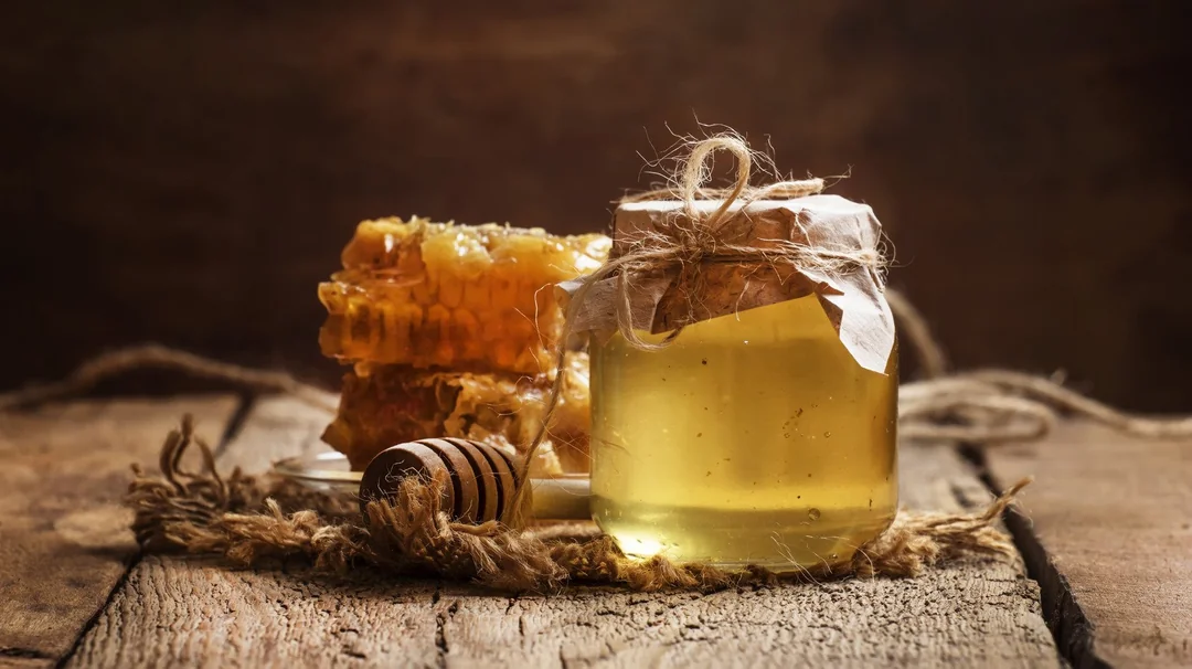 A jar of honey covered with paper and twine sits on a rustic wooden surface, next to honeycomb pieces and a wooden honey dipper. Warm, natural light highlights the golden color of the honey.