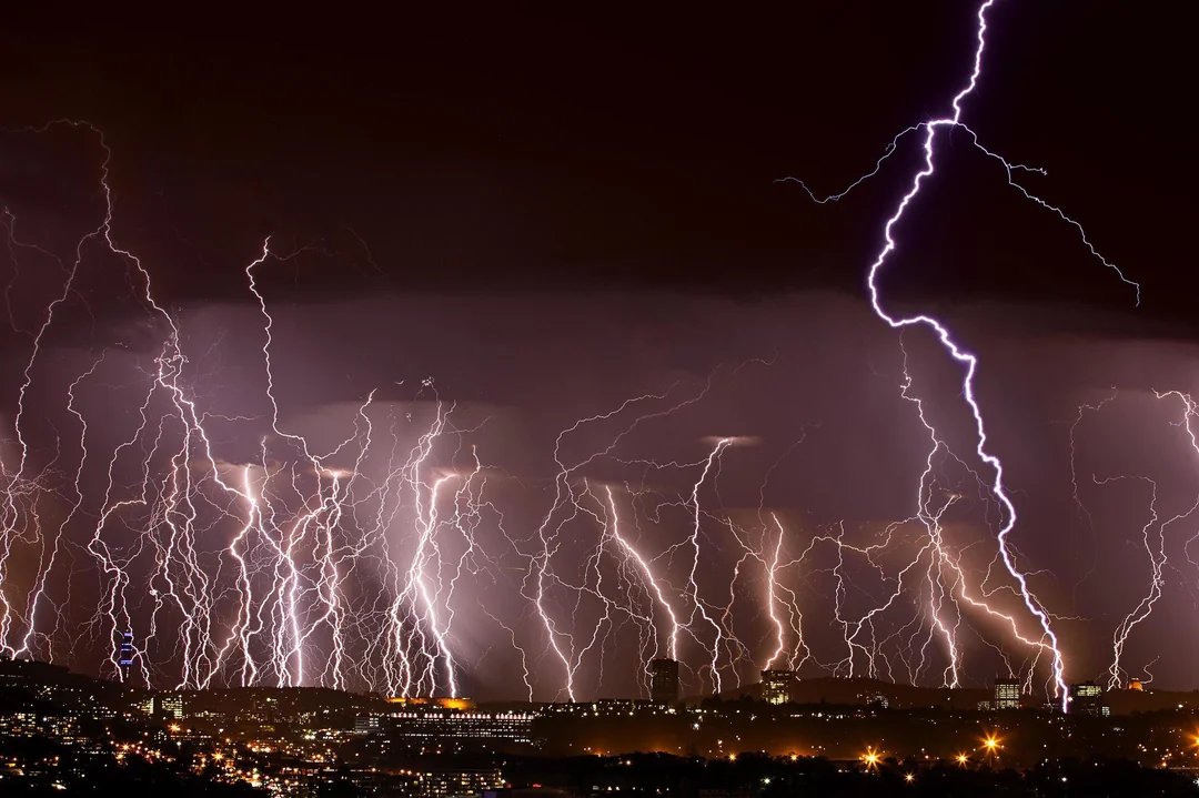 Multiple bolts of lightning dramatically illuminate the night sky above a city skyline, with bright flashes striking both clouds and the ground, creating a striking and intense scene.