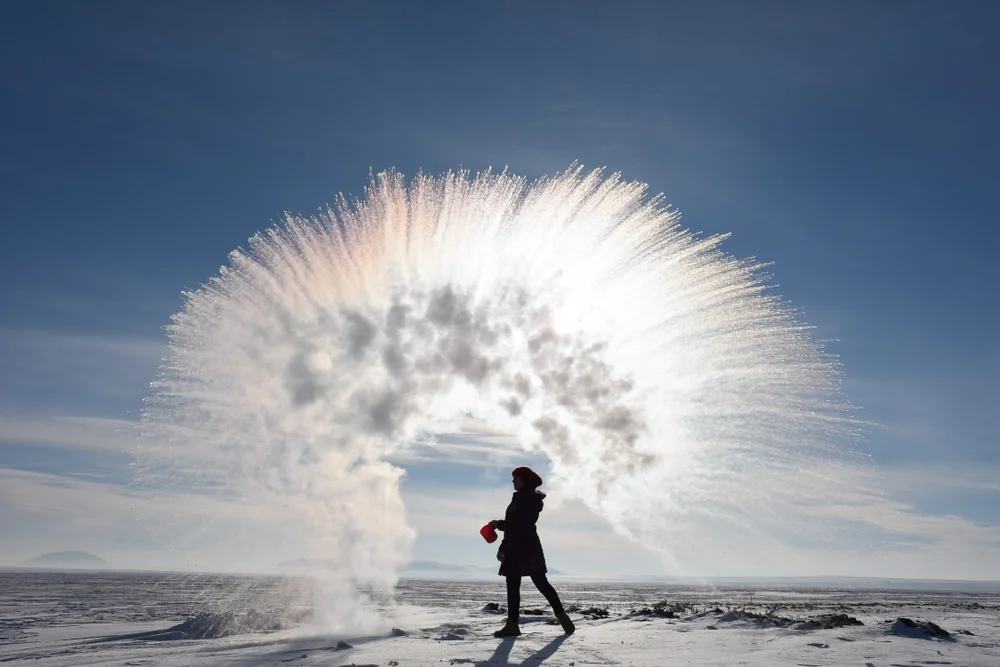 A person throws hot water into cold air, creating an arc of mist and ice crystals that fans out dramatically against a clear, blue sky on a snowy landscape.