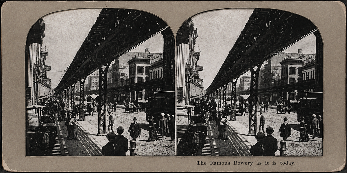 A black-and-white stereograph of New York City’s Bowery shows people walking under an elevated train track, with horse-drawn carriages and buildings lining the busy street.