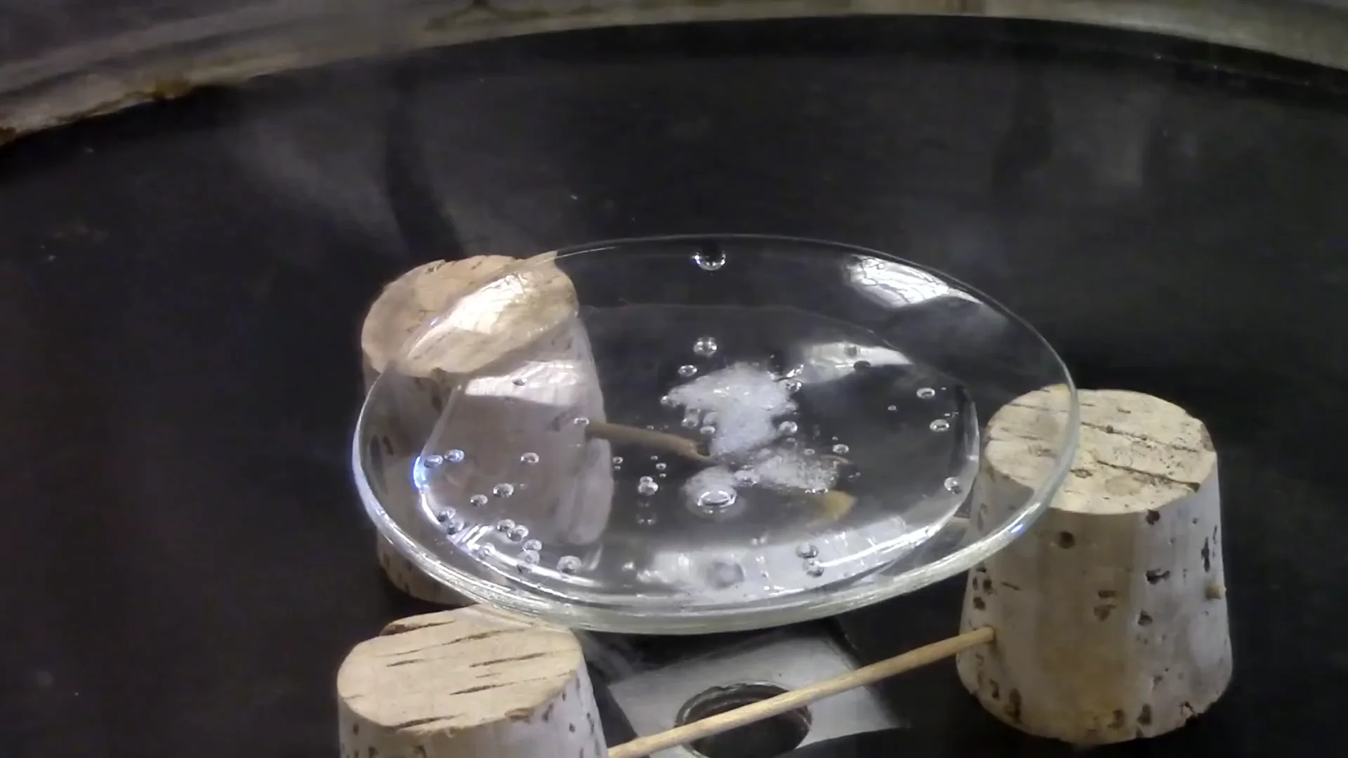 A glass dish with ice and water sits on three cork supports inside a dark chamber. Water droplets and bubbles are visible, and a wooden stick intersects under the dish through the corks.
