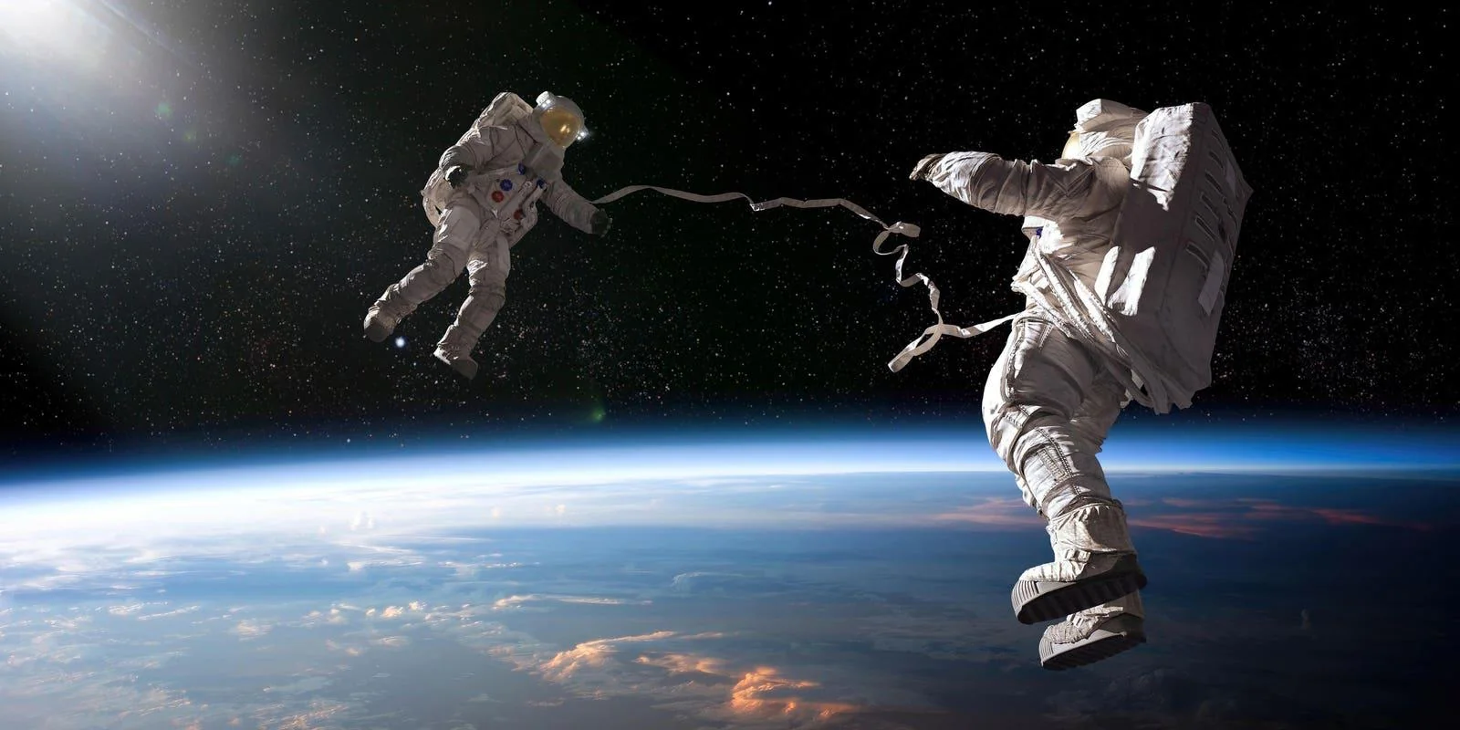 Two astronauts in spacesuits float above Earth, connected by a tether, with the planet’s curved horizon and a star-filled space background visible. Sunlight shines from the upper left corner.