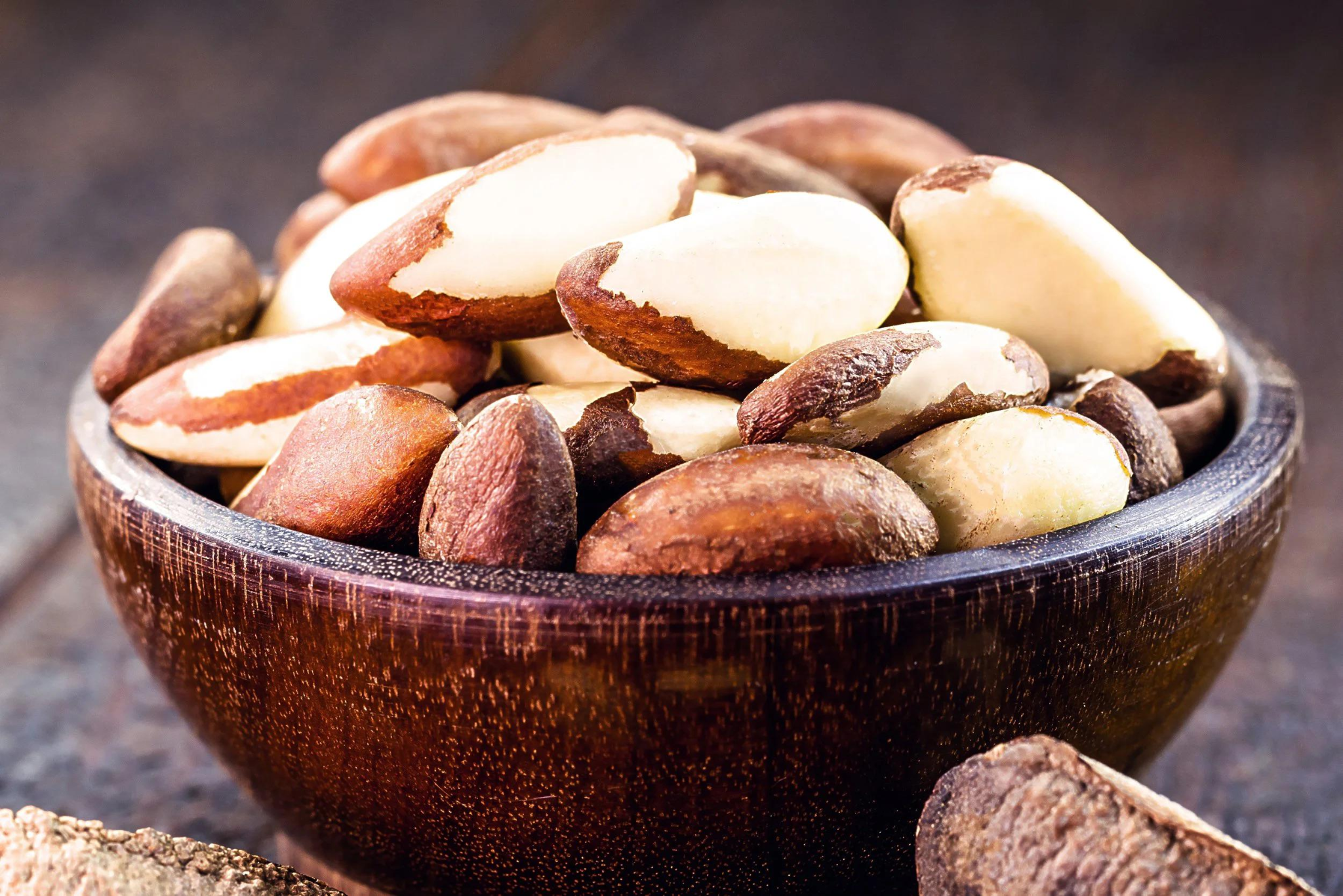 A wooden bowl filled with unshelled Brazil nuts sits on a wooden surface, with several nuts spilling over the edge.