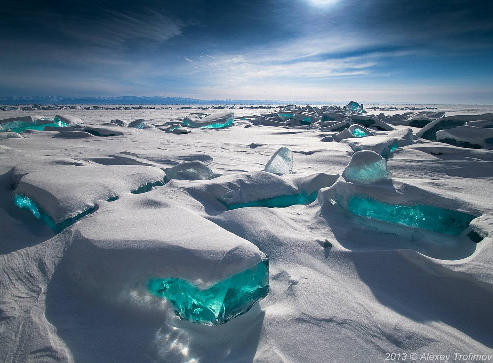 Large slabs of turquoise ice partially covered with snow stretch across a frozen landscape under a bright, cloudy sky. Sunlight glows on the icy surfaces, creating a surreal and striking winter scene.