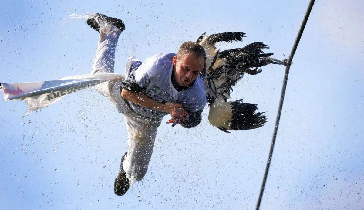 A man in midair splashes through water after slipping or falling, wearing white clothing, with droplets and water spray surrounding him against a blue sky.