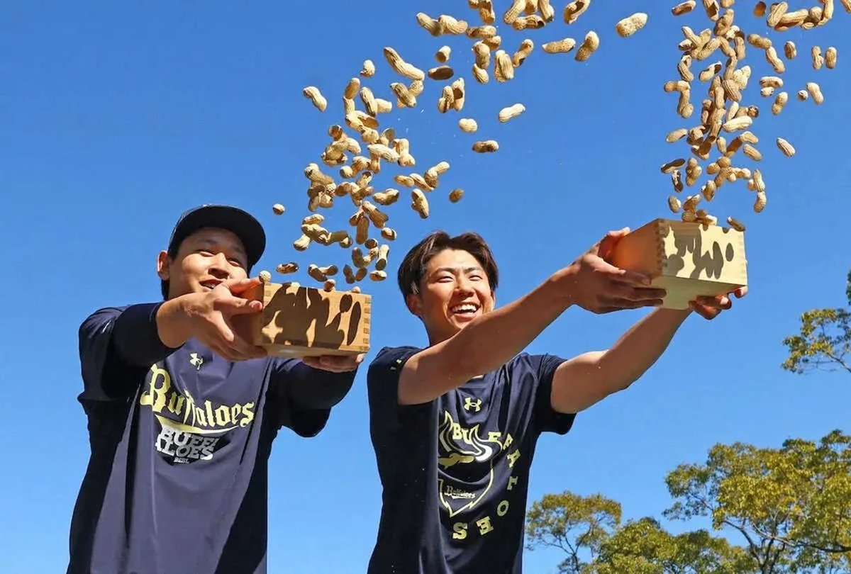 Two smiling young men in dark shirts toss peanuts from wooden boxes into the air outdoors on a sunny day, with a clear blue sky and some trees in the background.
