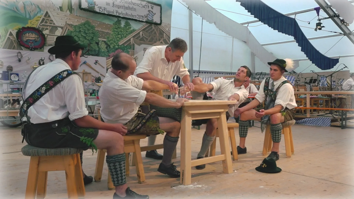 Five men in traditional Bavarian clothing sit on stools around a wooden table inside a decorated tent, engaged in an activity, possibly a competition or demonstration, with one man holding another’s arm.