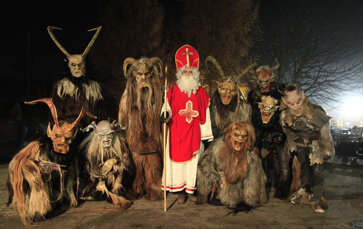 A person dressed as St. Nicholas stands in the center, surrounded by people in elaborate, scary Krampus costumes with horns and fur, posing outdoors at night.