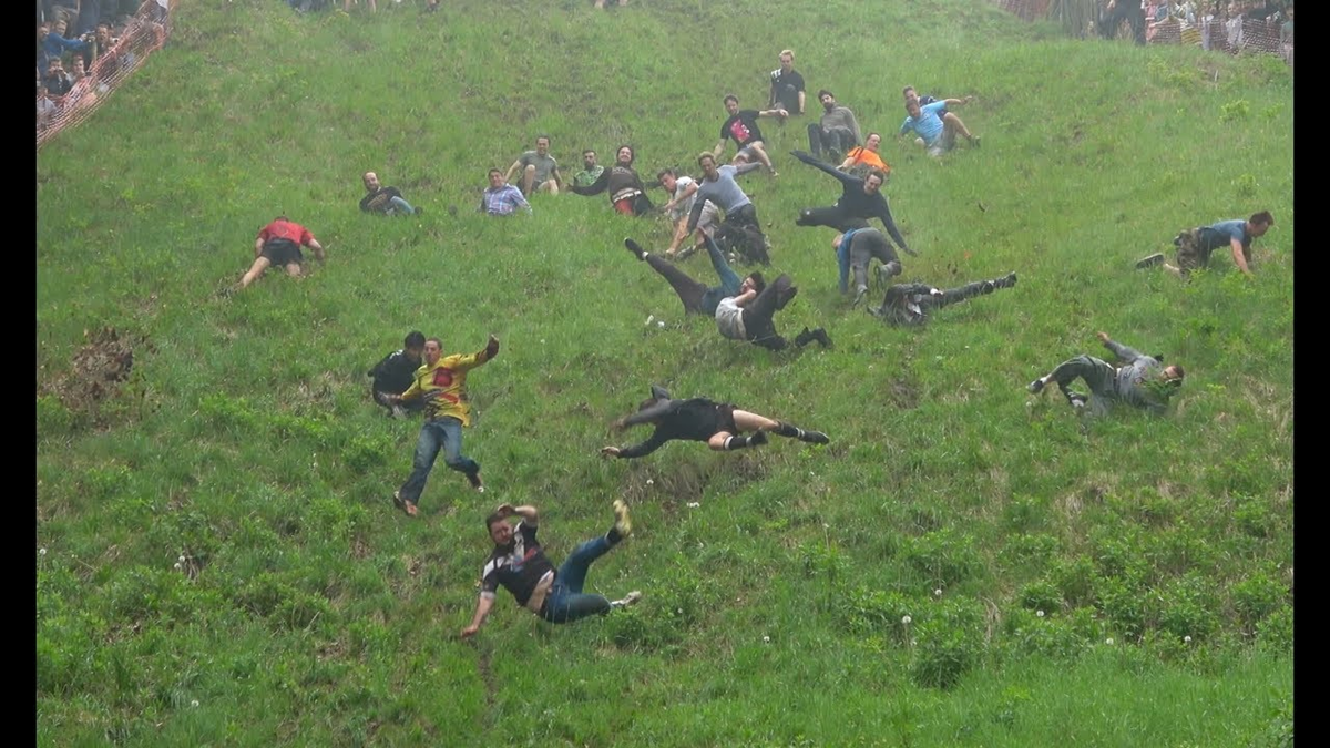 A group of people tumble and slide down a steep, grassy hill, some with arms and legs flailing, during a lively outdoor event or competition. Traditions.