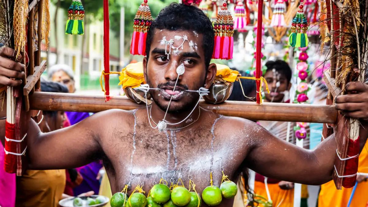 A man with limes pierced to his chest and face participates in a religious ritual, carrying a decorated wooden structure, surrounded by people and colorful decorations.
