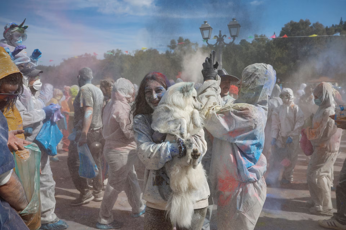 A crowd of people wearing protective clothing celebrate a colorful festival outdoors, throwing powder into the air. In the foreground, a smiling person holds a fluffy white dog, both covered in colorful powder.