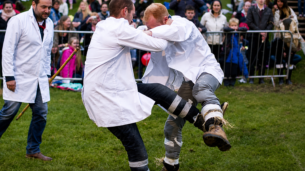 Two men wearing white coats and heavy boots grapple and kick each other in a grassy area, while a crowd watches from behind a metal barrier at an outdoor event. Another person in a coat stands nearby.
