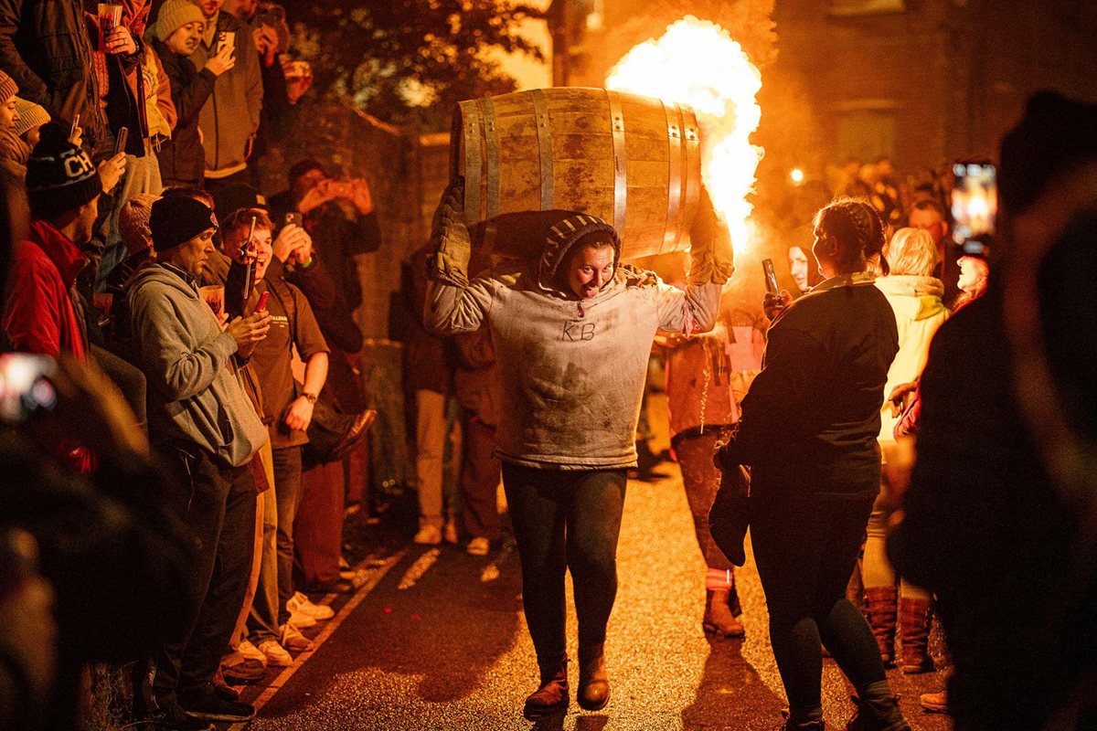 A person carries a flaming wooden barrel on their shoulders while walking down a street lined with cheering onlookers, some taking photos, during a nighttime event.