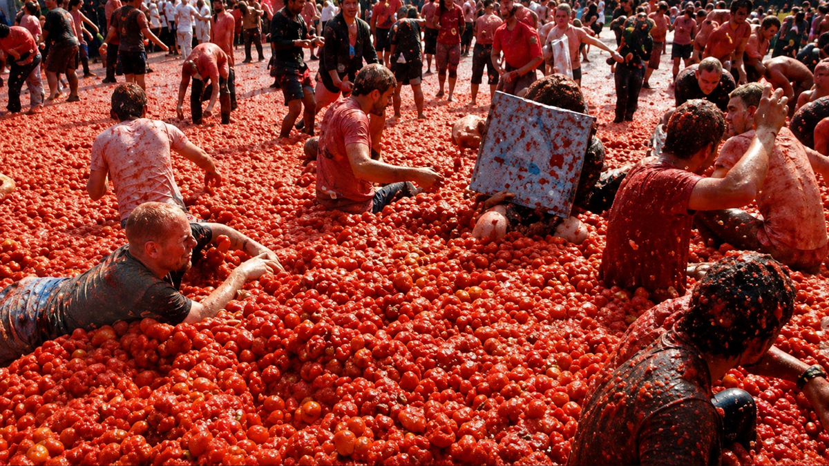 A large crowd of people covered in smashed tomatoes participate in a tomato fight festival, standing and crawling on a ground completely covered with red tomatoes and tomato pulp.