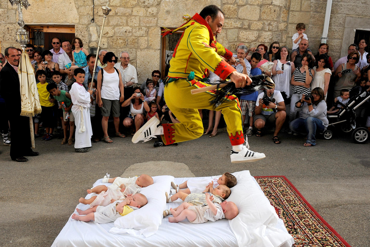 A man in a colorful costume jumps over several babies lying on a mattress outdoors, surrounded by a crowd of onlookers taking photos and watching the event.