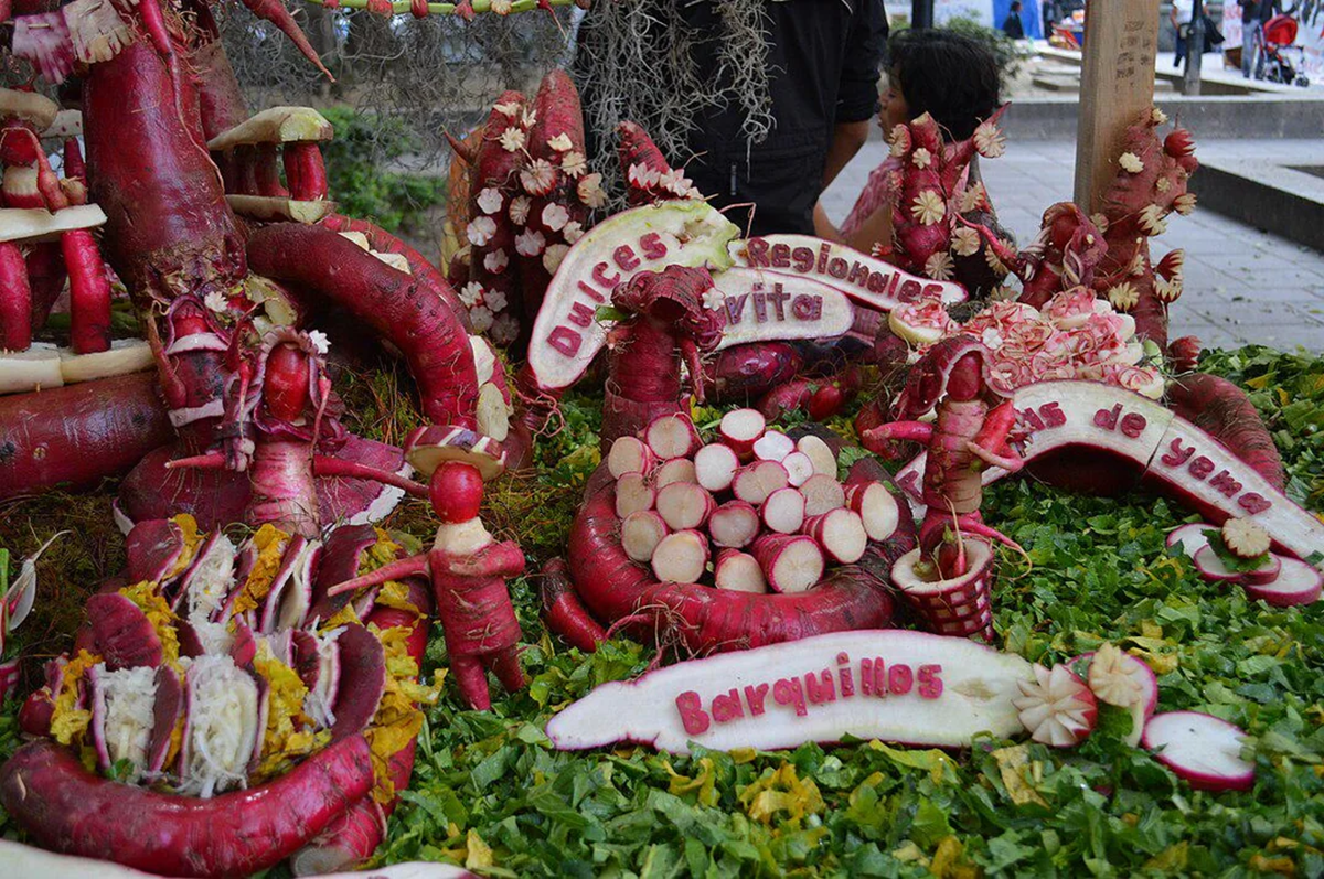 Colorful figurines, animals, and signs are intricately carved from reddish-purple radishes and displayed on a leafy green base at an outdoor event. People are visible in the background.