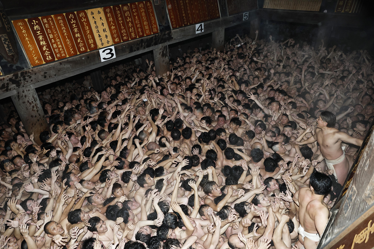 A dense crowd of men wearing white loincloths reaches upward inside a wooden hall, participating in a traditional Japanese festival. Numbered plaques and wooden tablets with Japanese writing hang on the walls above them.
