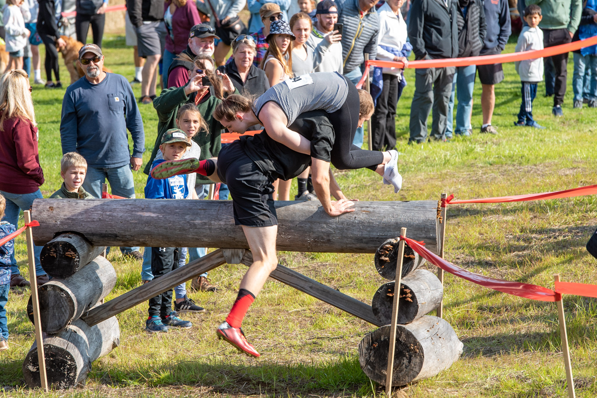 A man carrying a woman on his back climbs over a log obstacle during a race, while a crowd of spectators watches and takes photos.