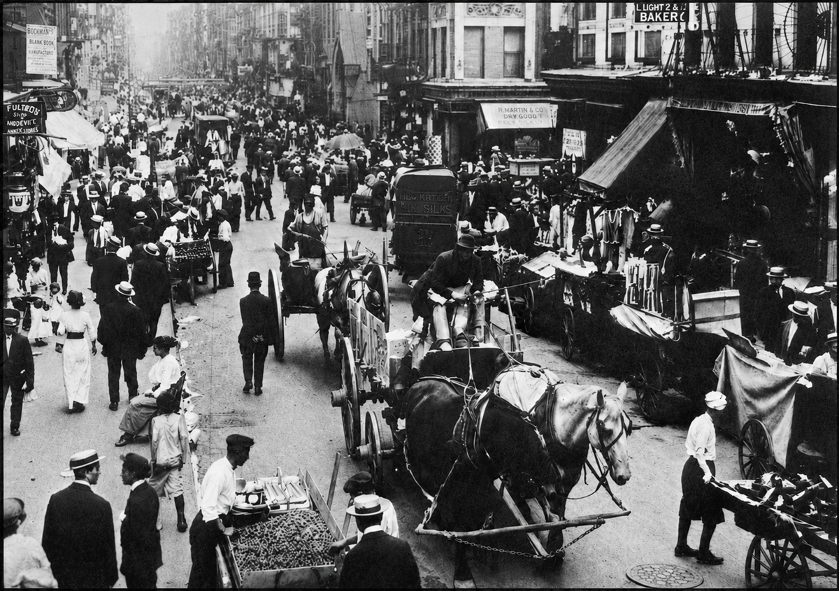 A busy city street in the early 1900s with many people walking, horse-drawn carriages, street vendors, and market stalls; buildings line both sides of the bustling urban scene.