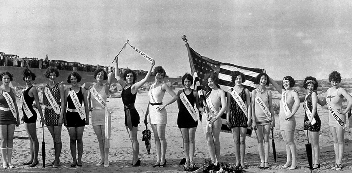 A black-and-white photo shows a group of women in swimsuits and sashes standing in a line on a beach. One holds an American flag. The background includes a crowd and cars. The image appears to be from the early 20th century.