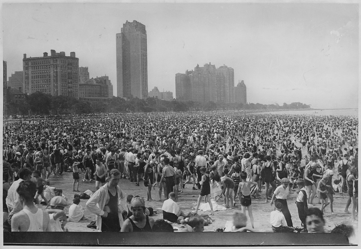 A historic black-and-white photo of a crowded beach with hundreds of people in vintage swimwear, tall city buildings in the background, and a hazy sky overhead.