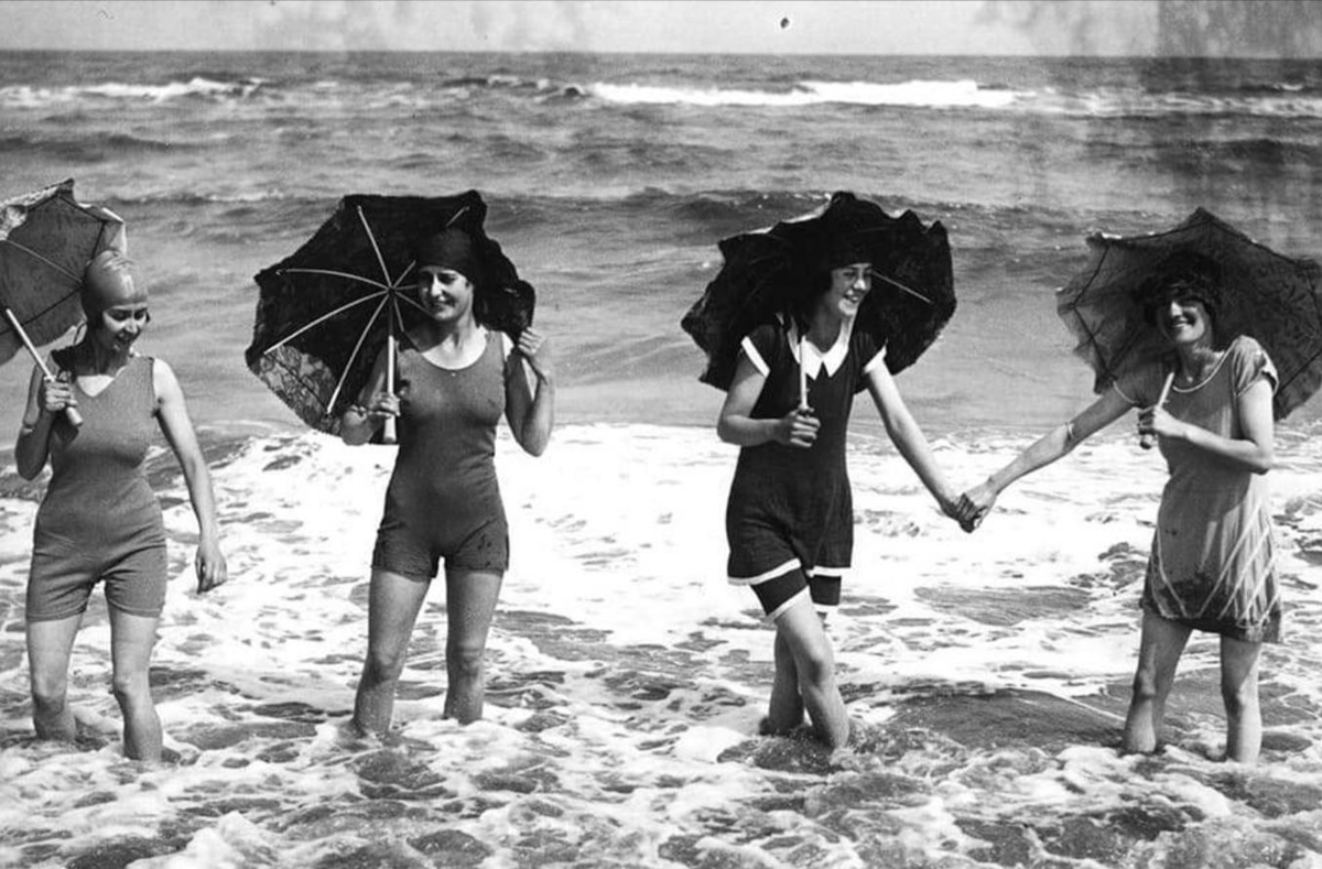 Four women in vintage swimsuits stand ankle-deep in the ocean, each holding an umbrella. Two of them are holding hands and smiling. The waves are gently washing over their feet.